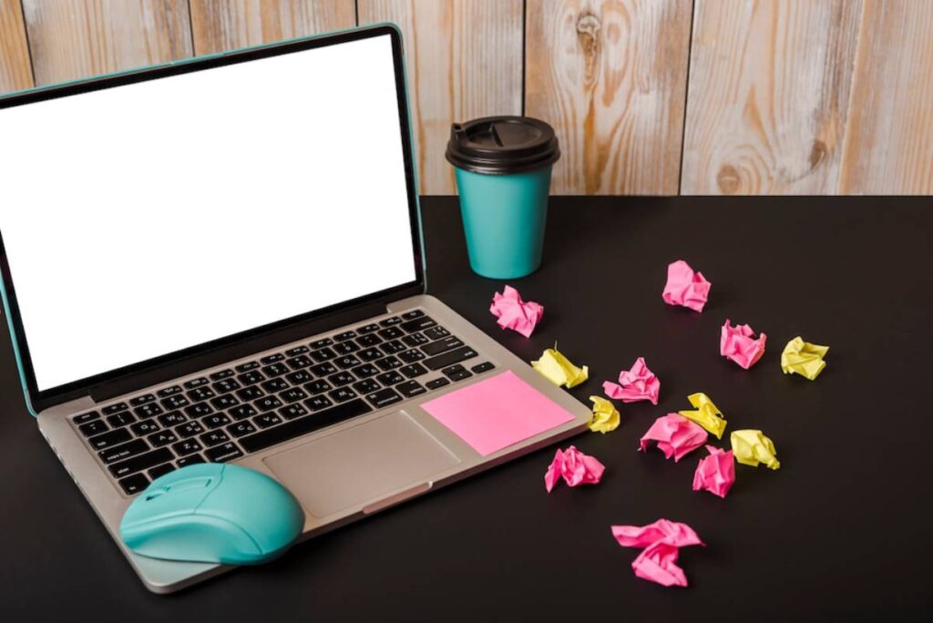 A laptop with a blank screen, a teal mouse, a coffee cup, and crumpled colorful papers scattered on a black desk.