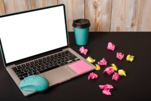 A laptop with a blank screen, a teal mouse, a coffee cup, and crumpled colorful papers scattered on a black desk.