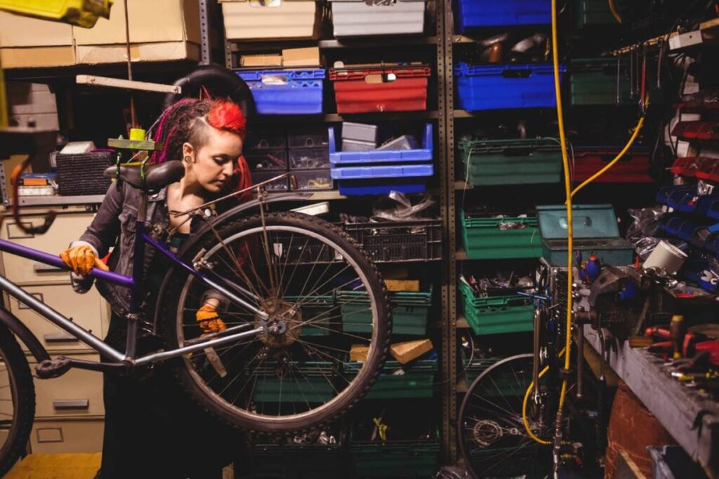 A person holds a bicycle in a cluttered workshop surrounded by colorful storage bins and various tools.