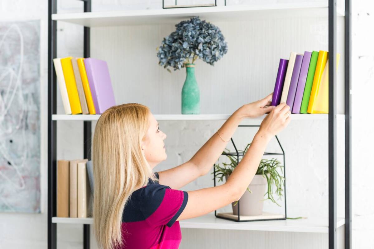 A woman arranges colorful books on a modern shelf, alongside a potted plant and a decorative vase with flowers.