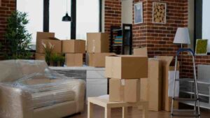 A cozy, packed living room filled with cardboard boxes and a wrapped sofa, surrounded by plants and warm brick walls.