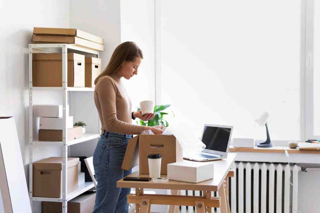 A woman stands at a desk, organizing items, with cardboard boxes, a laptop, and a potted plant in a bright, minimalist workspace.