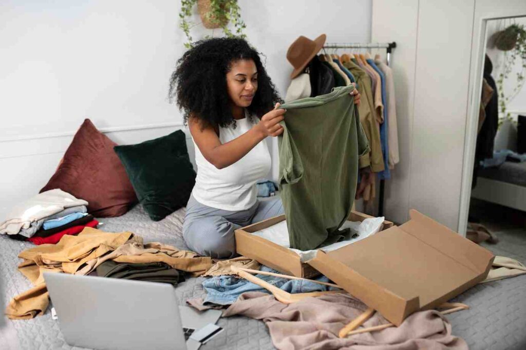 A person seated on a bed examines a green T-shirt, surrounded by clothing and a laptop, in a cozy, styled room.