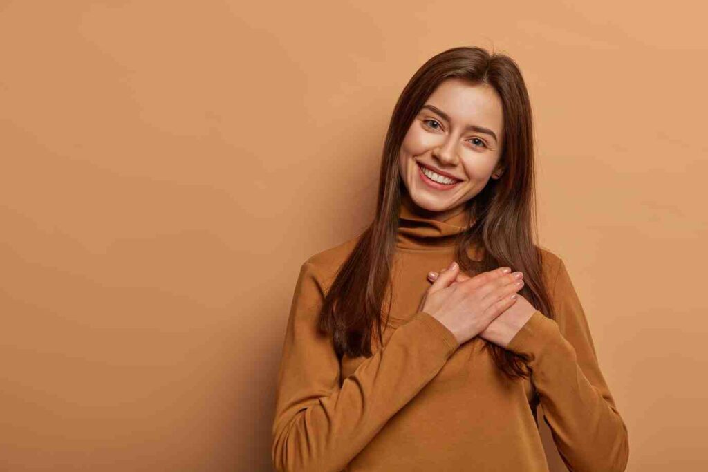 A woman in a brown turtleneck sweater stands with her hands clasped near her chest against a warm, peach-colored background.