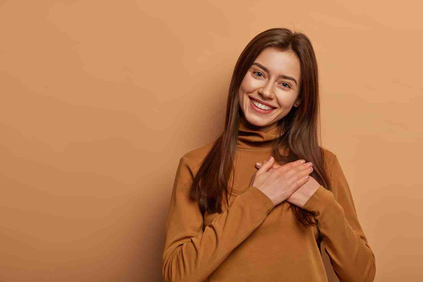 A woman in a brown turtleneck sweater stands with her hands clasped near her chest against a warm, peach-colored background.