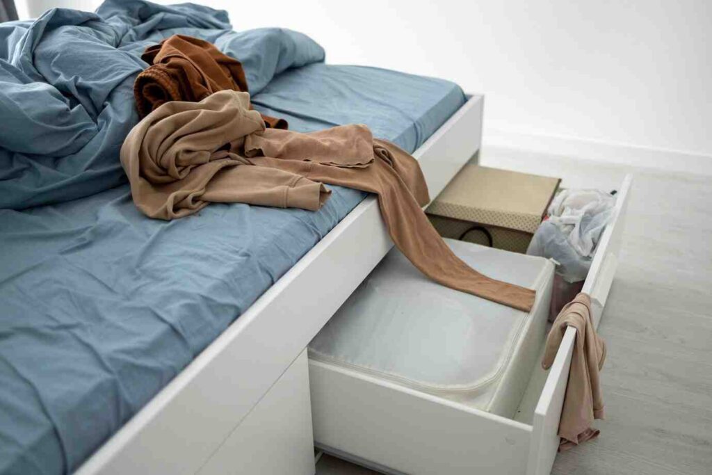 A messy bed with crumpled blue sheets and various clothing items piled on top, alongside an open drawer revealing storage and trash bags.