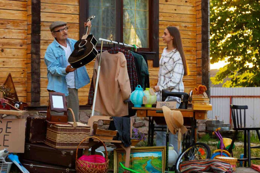 A man holds a black guitar while a woman stands nearby, surrounded by items for sale at a yard sale outside a wooden house.