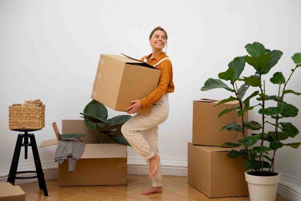 A person in an orange shirt and beige pants balances on one leg while carrying a large moving box amidst packed boxes and plants.