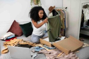 A woman examines a green shirt while surrounded by neatly folded clothes and an open box on her bed, with a laptop nearby.
