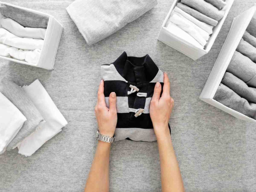A person's hands hold a neatly folded black and white shirt, surrounded by organized boxes of rolled gray and white clothing on a gray surface.