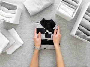A person's hands hold a neatly folded black and white shirt, surrounded by organized boxes of rolled gray and white clothing on a gray surface.