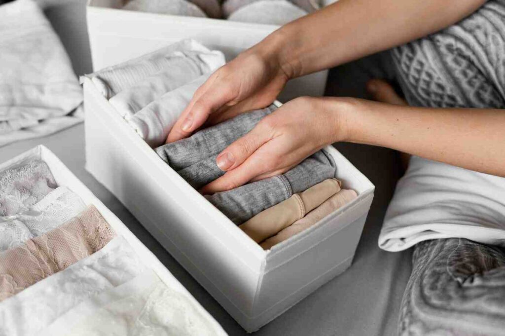 A person carefully organizes neatly folded clothes in a white storage box, with additional garments visible in the background.