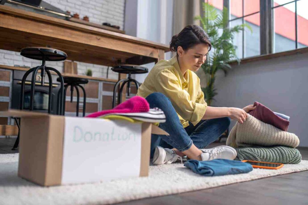 A person sits on a rug organizing clothes into a donation box surrounded by neatly folded items and a smartphone nearby.