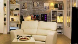 Cozy living room featuring a cream sofa, snacks on a table, a gaming setup, and decorative shelves against a wooden wall.