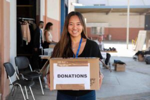 A woman holds a cardboard box labeled DONATIONS in a donation center, surrounded by people and clothing items.