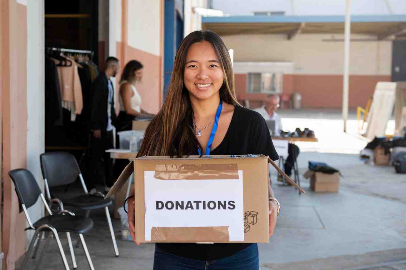 A woman holds a cardboard box labeled DONATIONS in a donation center, surrounded by people and clothing items.