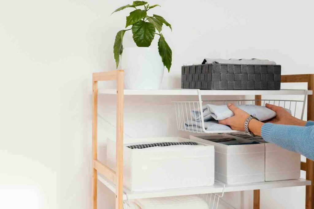 A person arranges folded clothes on a modern shelving unit with storage baskets and a potted plant on top.