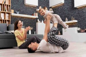 A parent lifts a joyful baby overhead while another parent sits nearby, clapping and smiling in a cozy living room.