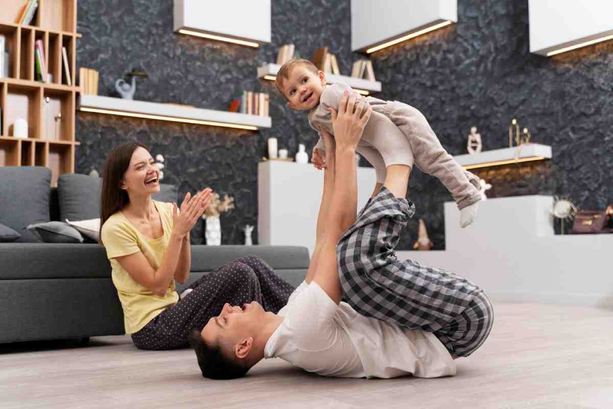 A parent lifts a joyful baby overhead while another parent sits nearby, clapping and smiling in a cozy living room.