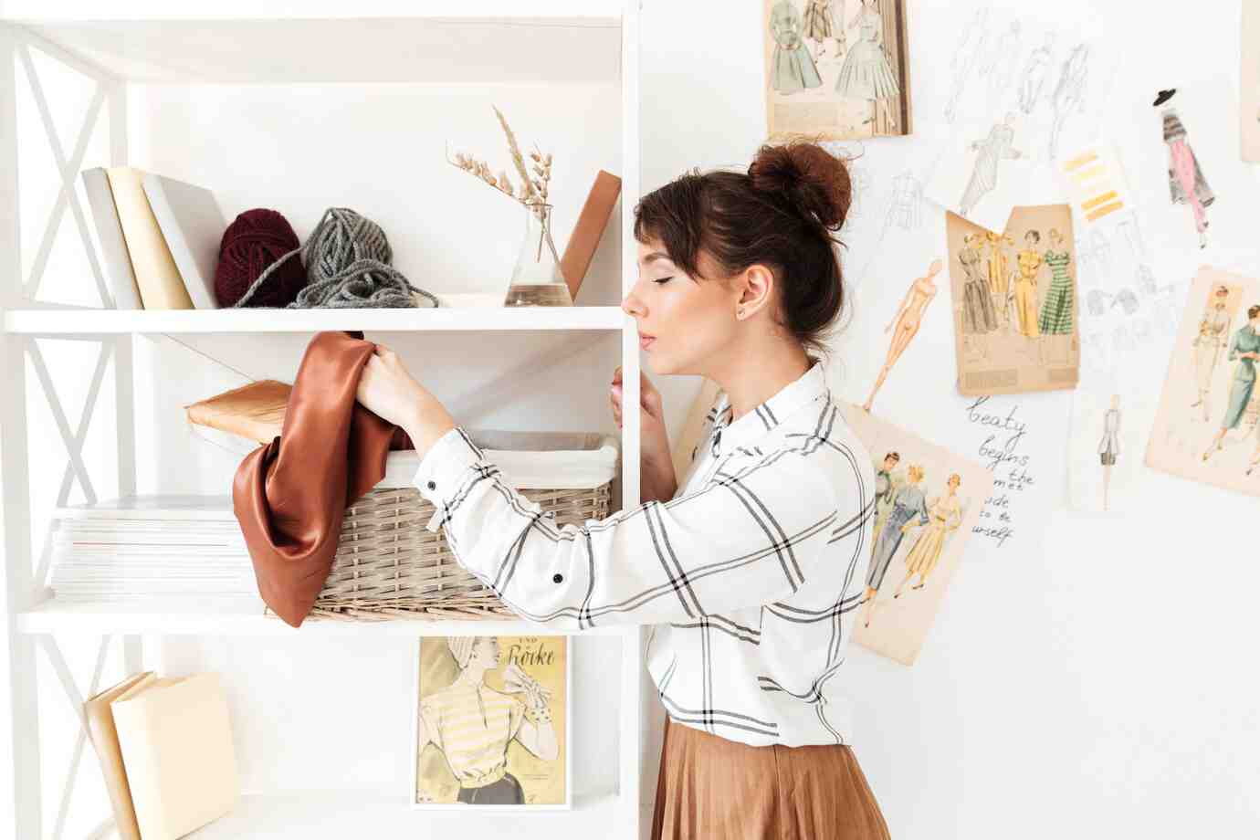 A person in a plaid shirt organizes a shelf with yarn, fabric, and books, surrounded by fashion sketches on the wall.