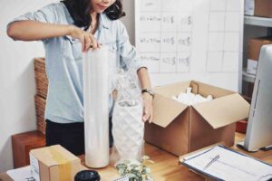 A woman unwraps a decorative vase while organizing packaging materials at a workspace filled with boxes and paperwork.