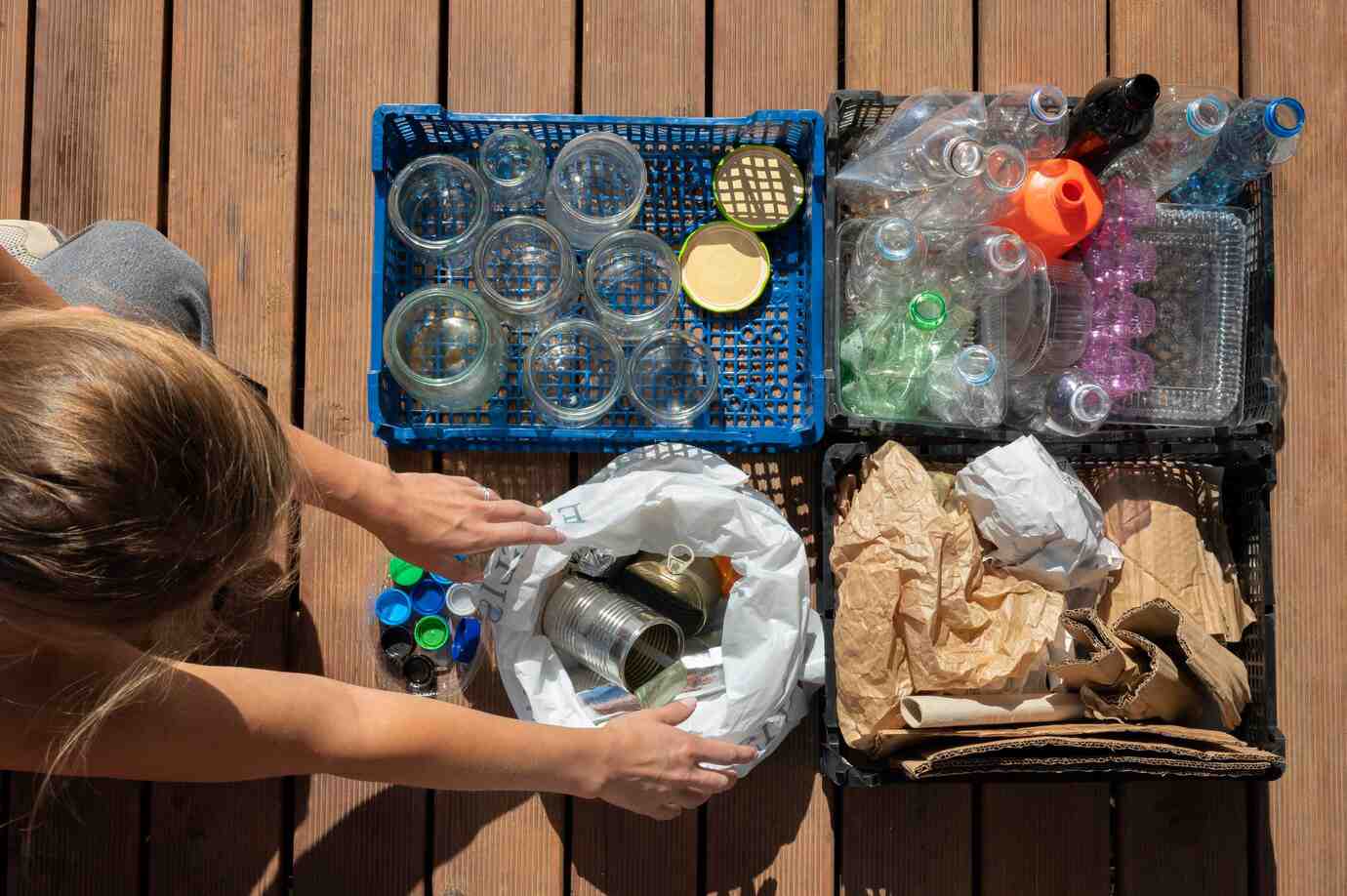 A person organizes recyclables, including glass jars, plastic bottles, and paper, on a wooden surface under bright sunlight.