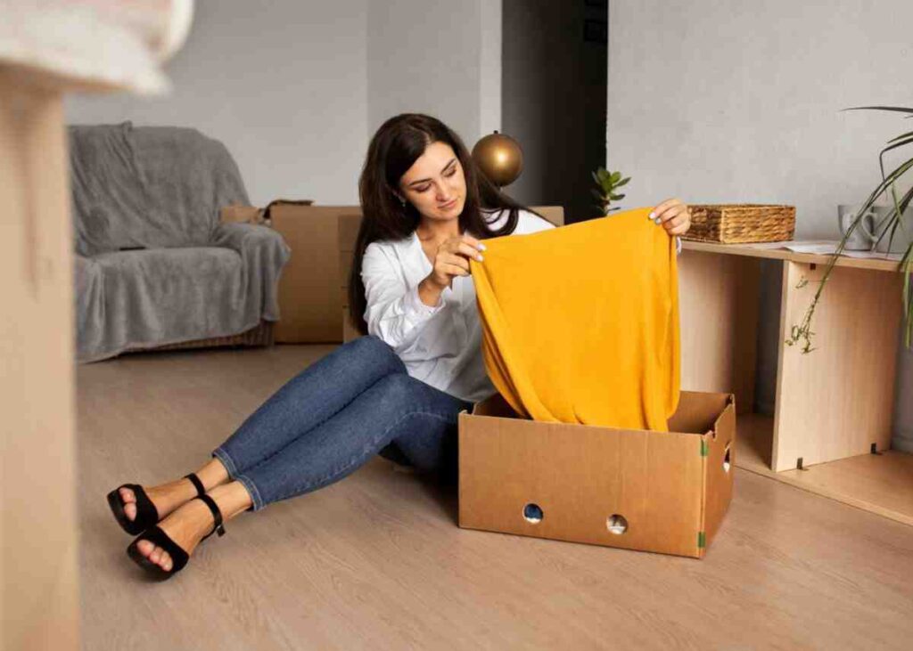 A person sits on the floor, holding a yellow garment, as they unpack a box in a cozy, modern living space.