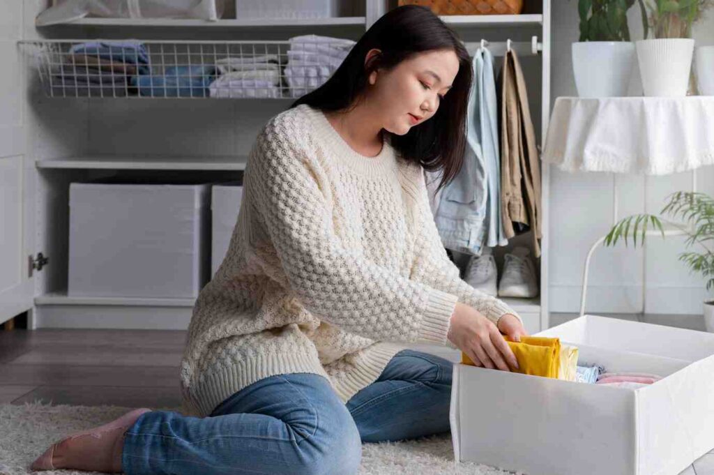 A person sits on a plush rug, organizing colorful clothes into a white storage box in a tidy, well-decorated room.