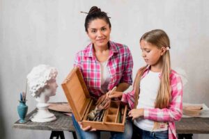 A woman and a girl in matching plaid shirts explore a wooden art box filled with tools, against a neutral backdrop with sculpting materials.