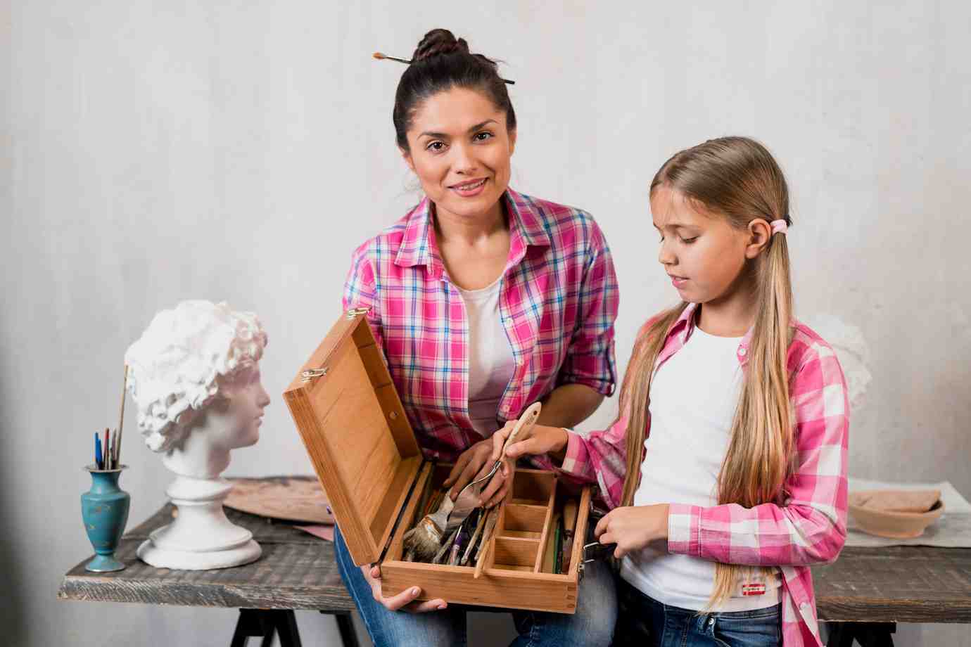 A woman and a girl in matching plaid shirts explore a wooden art box filled with tools, against a neutral backdrop with sculpting materials.