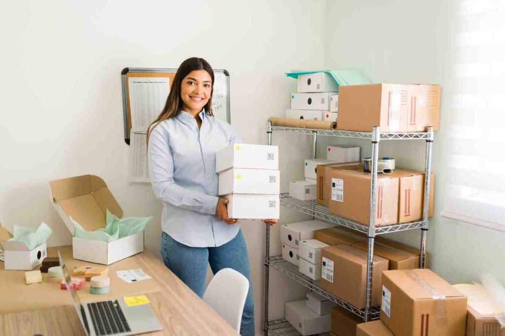 A woman holds stacked boxes in a workspace filled with organized packages and a laptop on a wooden desk.