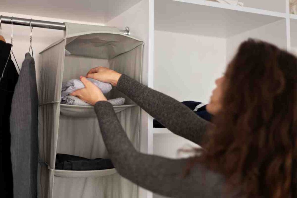A person organizes clothing in a fabric storage unit, folding towels while surrounded by hanging garments in a tidy closet space.