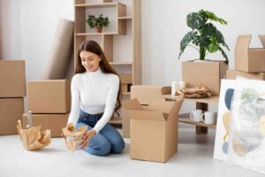 A woman kneels on a bright floor, unpacking items from cardboard boxes in a spacious, modern room with a plant and decorative artwork.