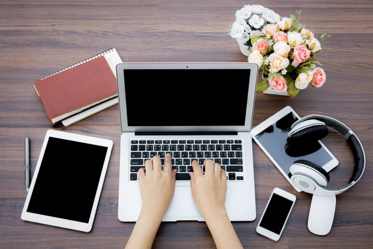 A top-down view of a workspace with a laptop, tablet, smartphone, headphones, a pen, notebooks, and a flower arrangement.