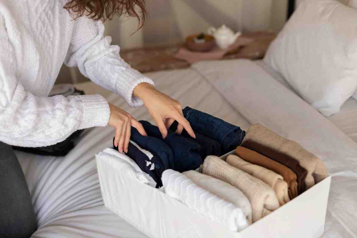 A person’s hands organize neatly rolled cozy sweaters in a white box, showcasing a variety of earth-toned fabrics.