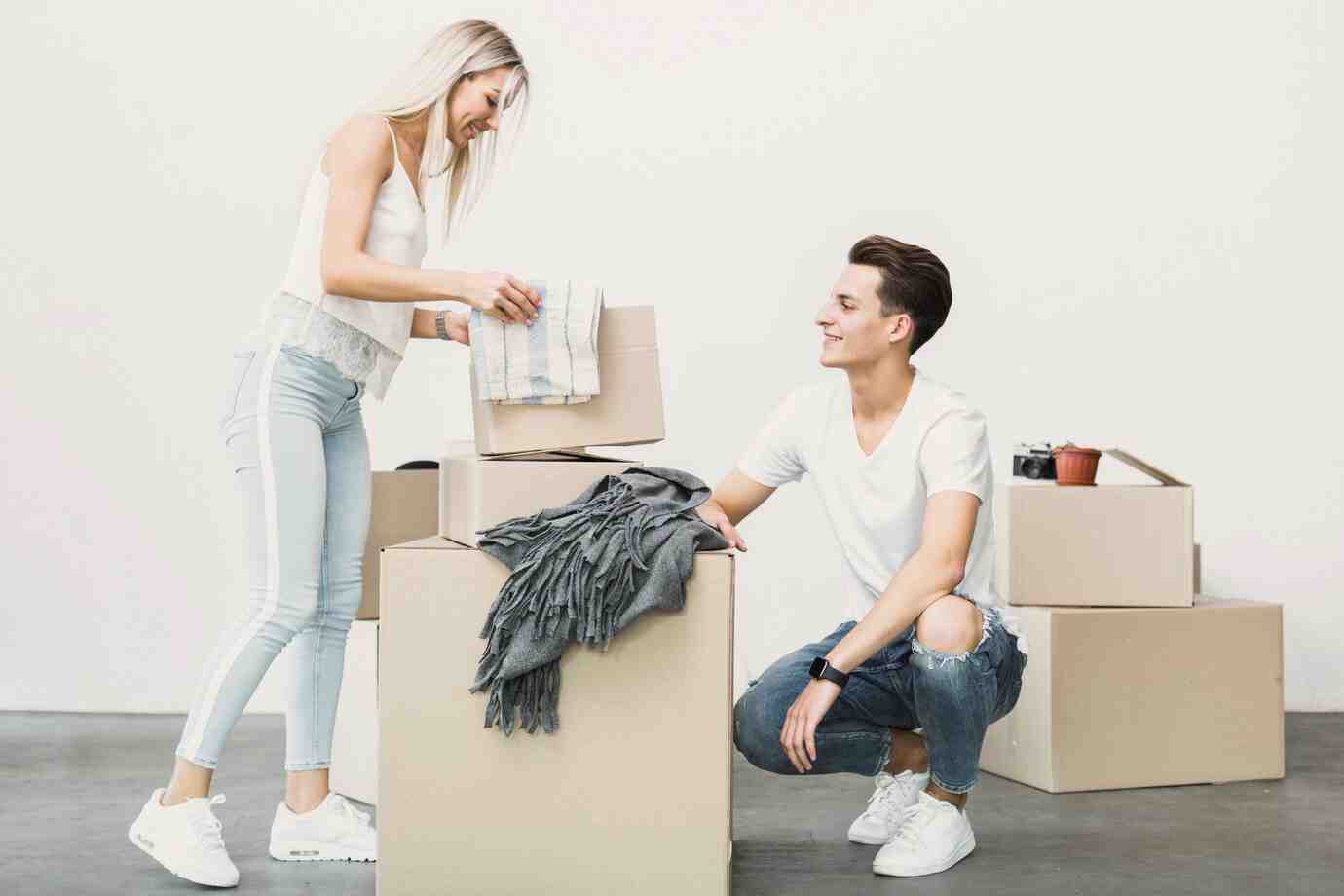 A young woman arranges items on top of cardboard boxes, while a young man attentive watches, in a minimalistic indoor setting.