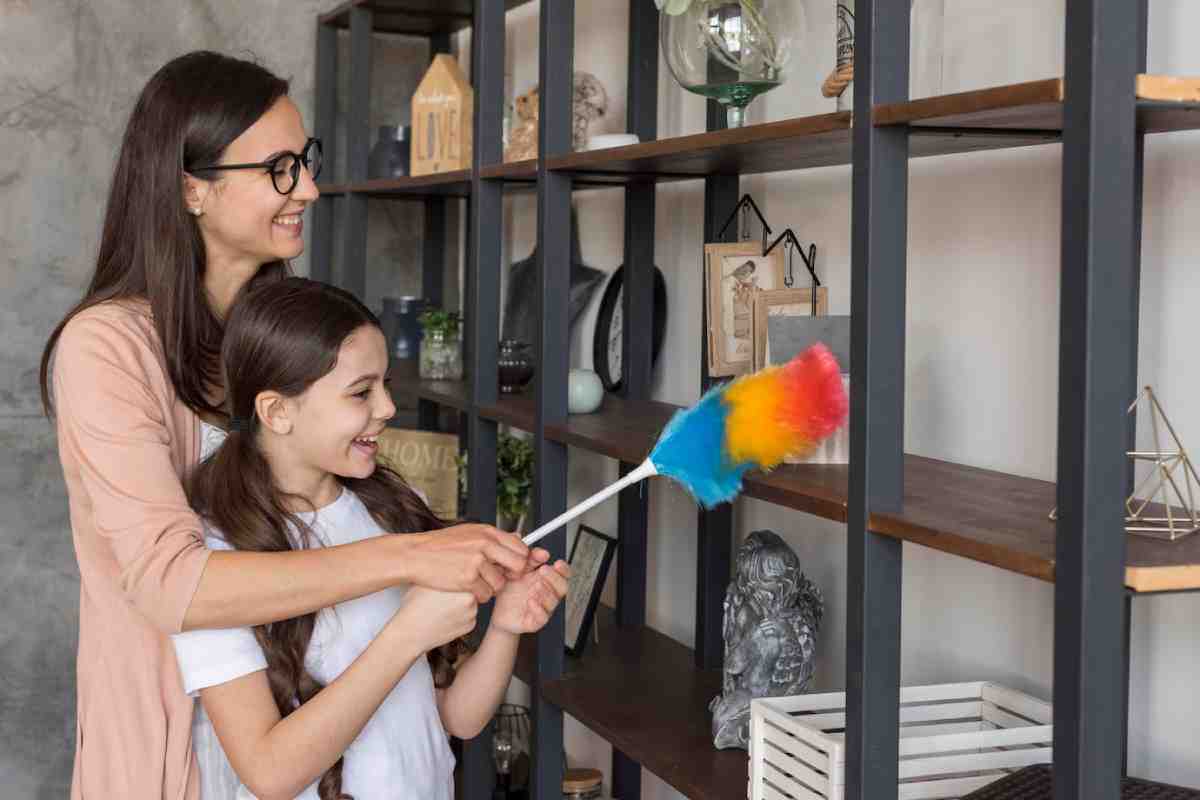 A woman guides a child in cleaning a decorative shelf with a colorful duster, surrounded by various ornaments and plants.