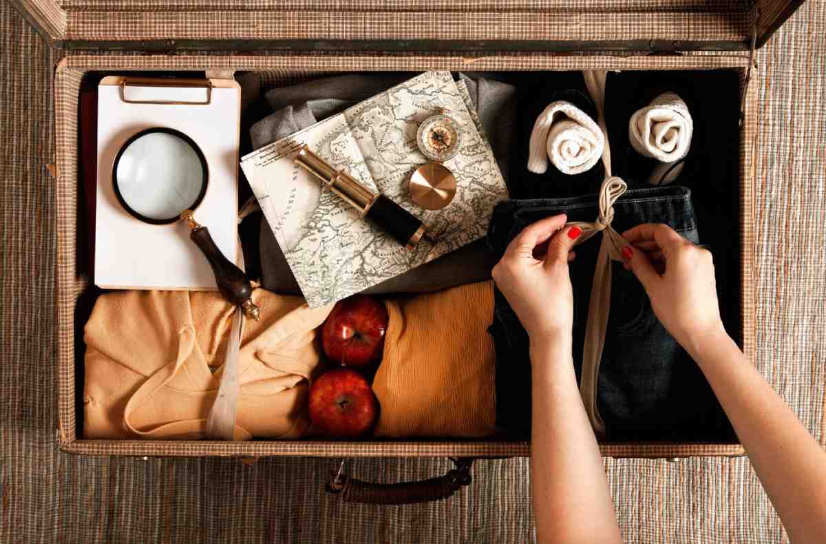A person's hands tie a ribbon around neatly packed clothes, a map, and travel essentials in a vintage suitcase.