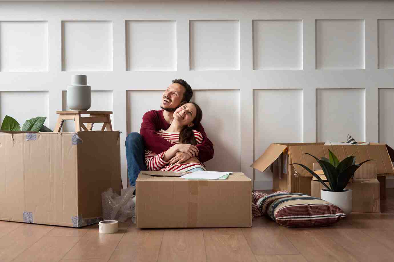 A couple sits closely together on the floor surrounded by unpacked cardboard boxes and a small potted plant in a cozy, new home.