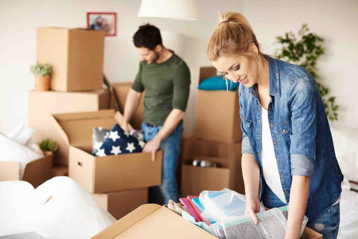 A woman in a denim shirt organizes colorful items in a box, while a man sorts items in the background among unpacked cardboard boxes.