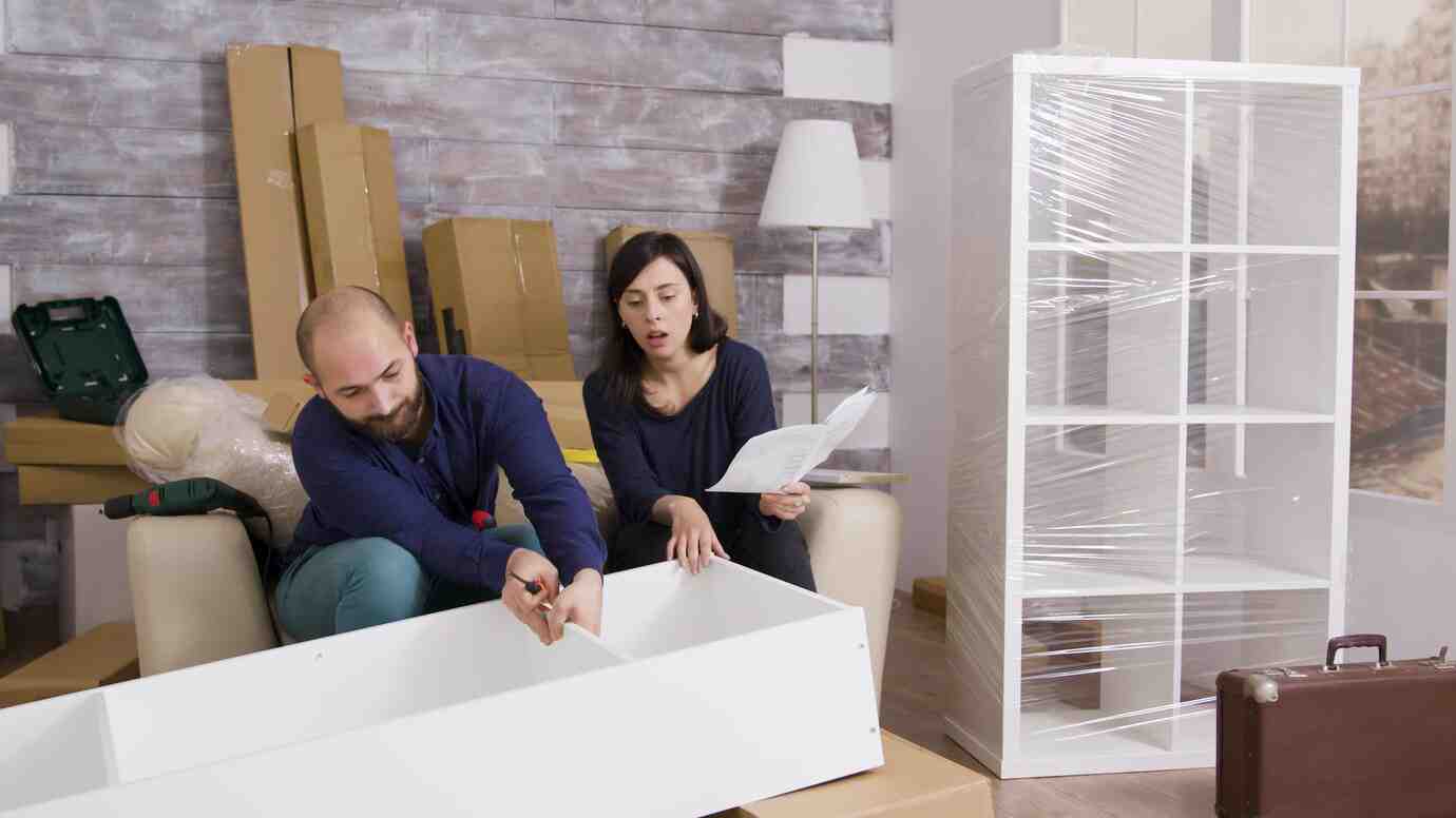 A couple sits on a couch, discussing assembly instructions for flat-packed furniture amid unpacked boxes and a new shelf wrapped in plastic.