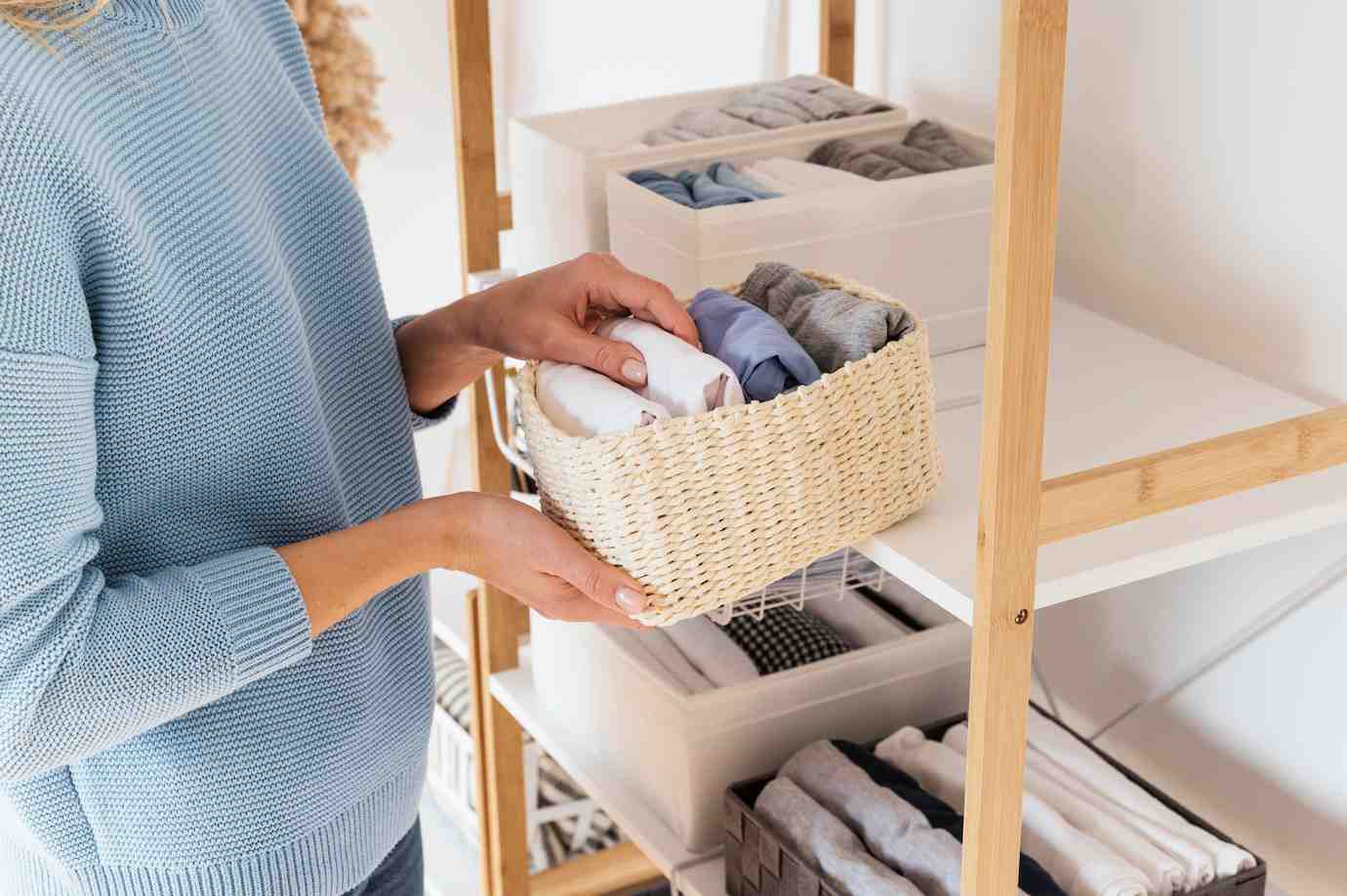 A woman in a blue sweater organizes folded clothes in a woven basket on a shelf, surrounded by neatly arranged containers.