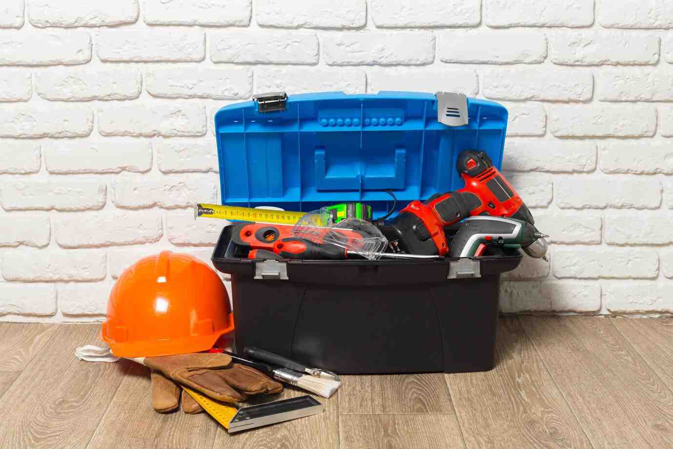 A blue toolbox filled with tools, including drills and a measuring tape, alongside an orange hard hat and work gloves on a wooden floor.