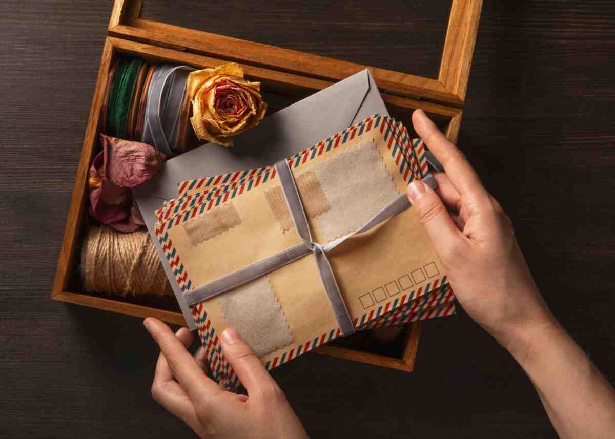 A hand holds vintage envelopes tied with ribbon above a wooden box containing ribbon, dried flowers, and twine.