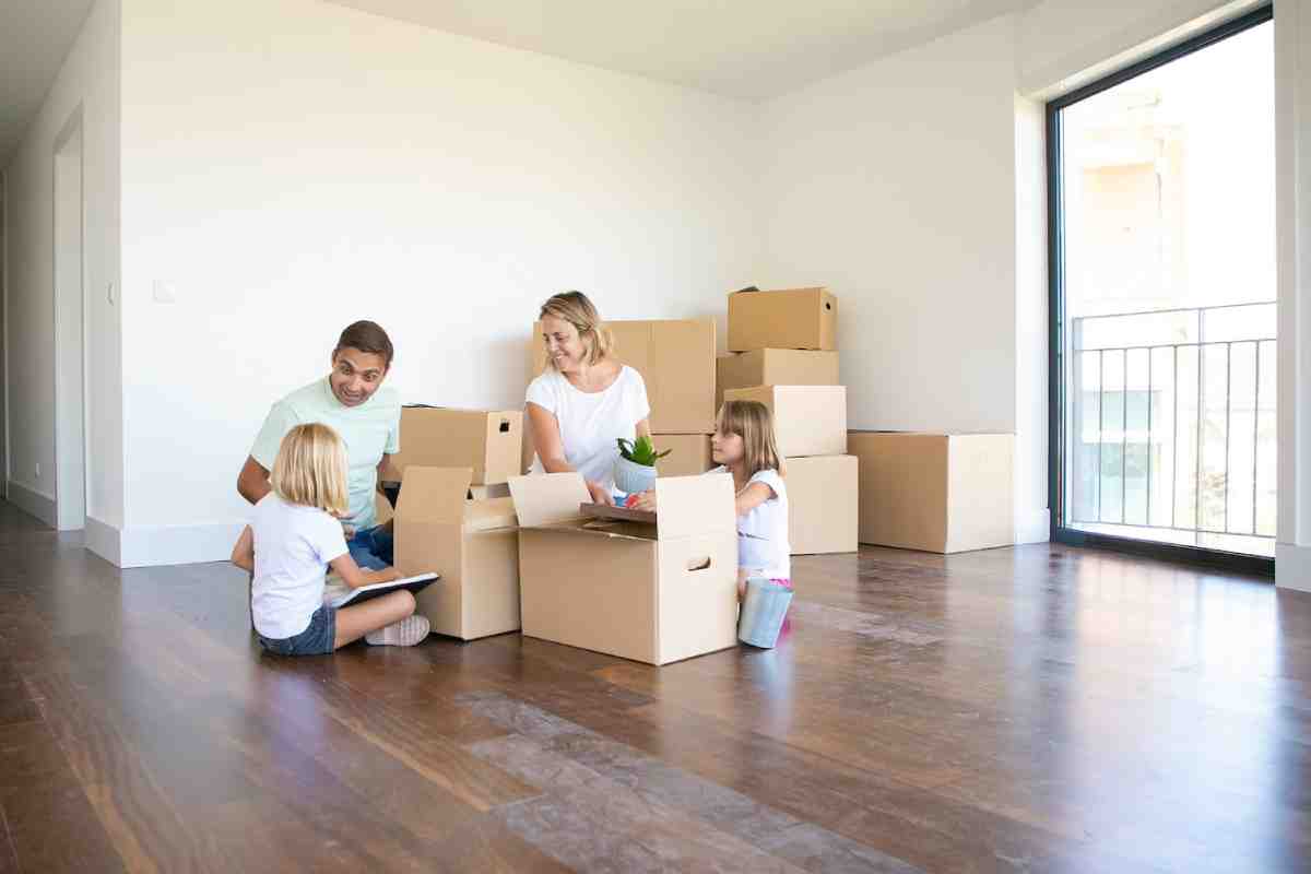 A family of four, including two children, unpacks moving boxes in a bright, empty room with wooden floors and a large window.