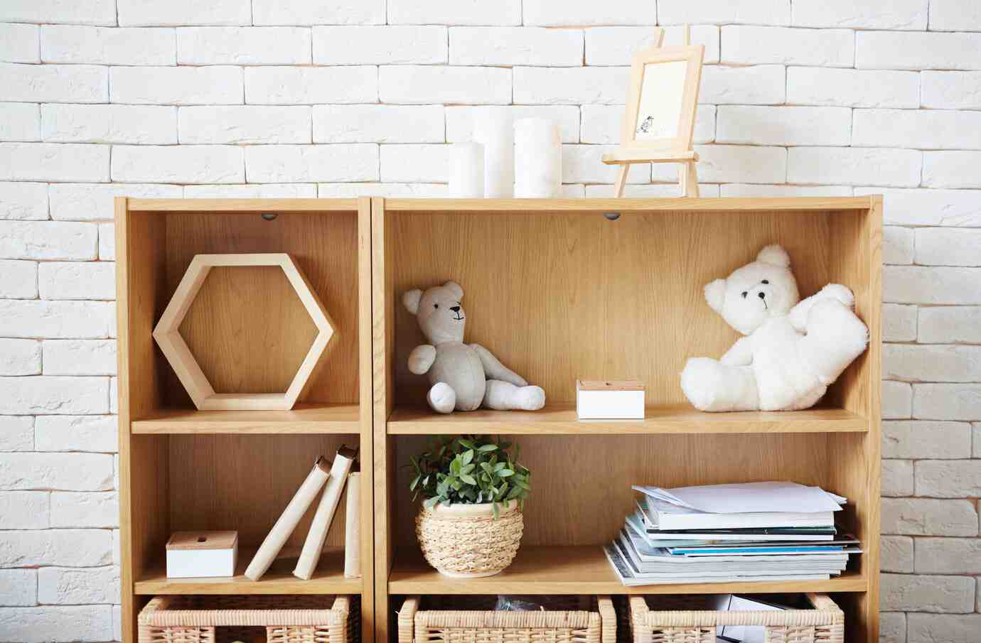 A wooden bookshelf with stuffed bears, books, a hexagonal frame, plants, and a small picture frame against a white brick wall.