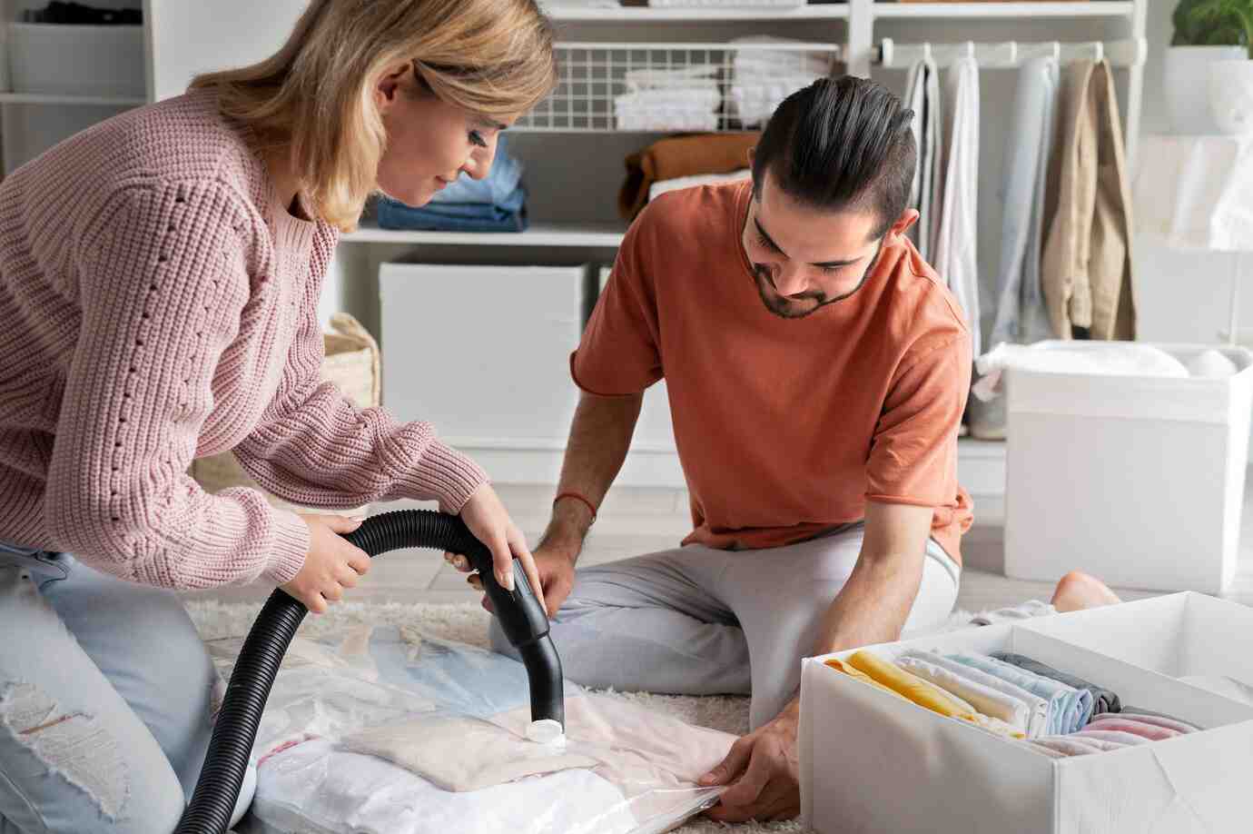 A woman uses a vacuum bag on clothes while a man assists, surrounded by neatly organized storage in a modern home setting.