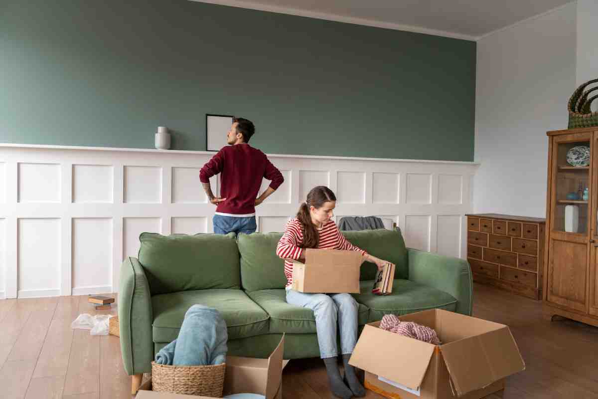A woman sits on a green couch unpacking boxes, while a man stands nearby, observing the room's decor and organization.