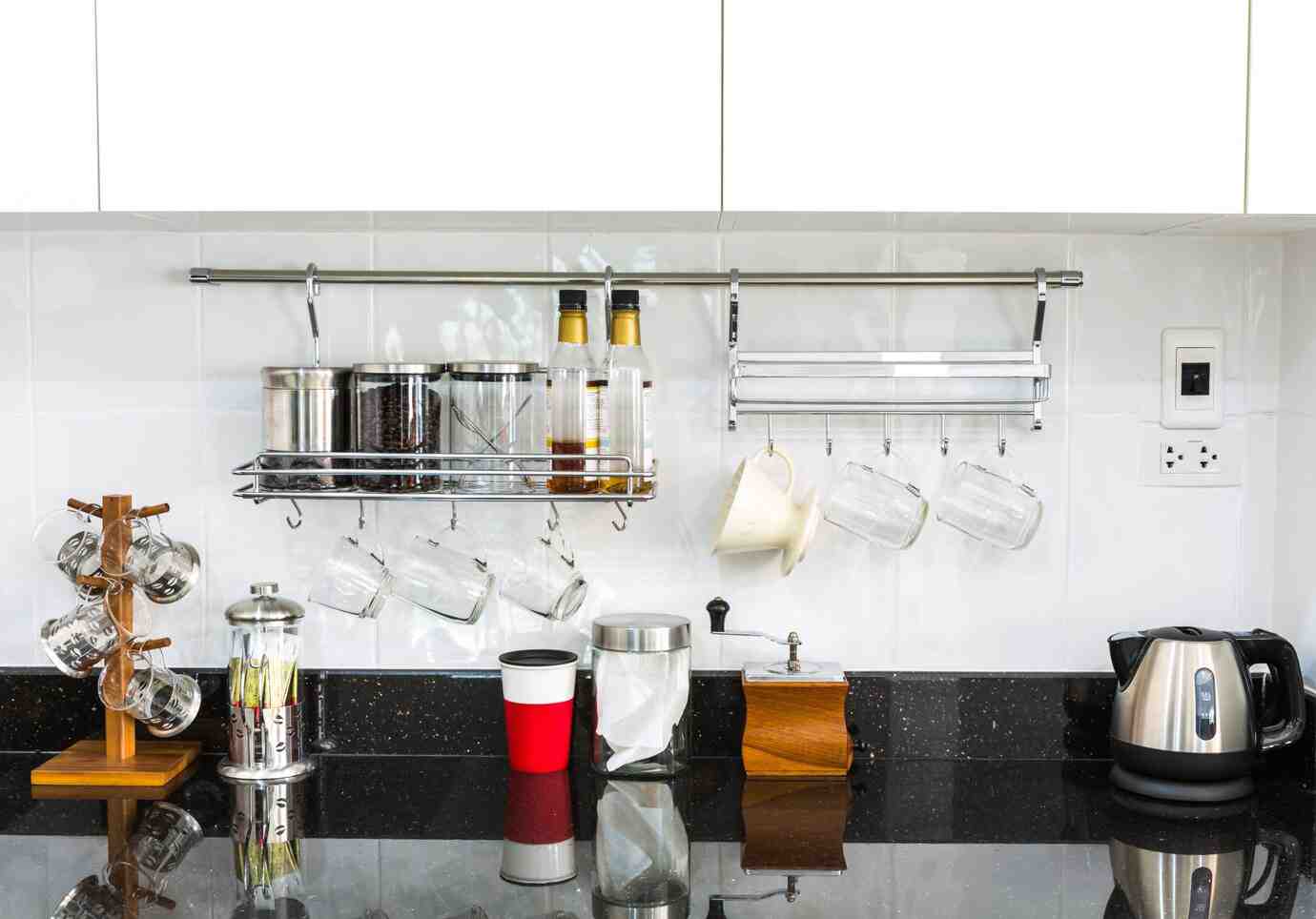 A modern kitchen shelf displaying jars, bottles, hanging mugs, and a kettle on a sleek black countertop.
