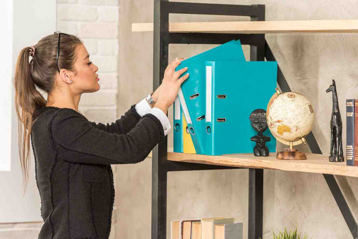 A woman organizes bright blue binders on a wooden shelf, accompanied by a globe, a small statue, and books.